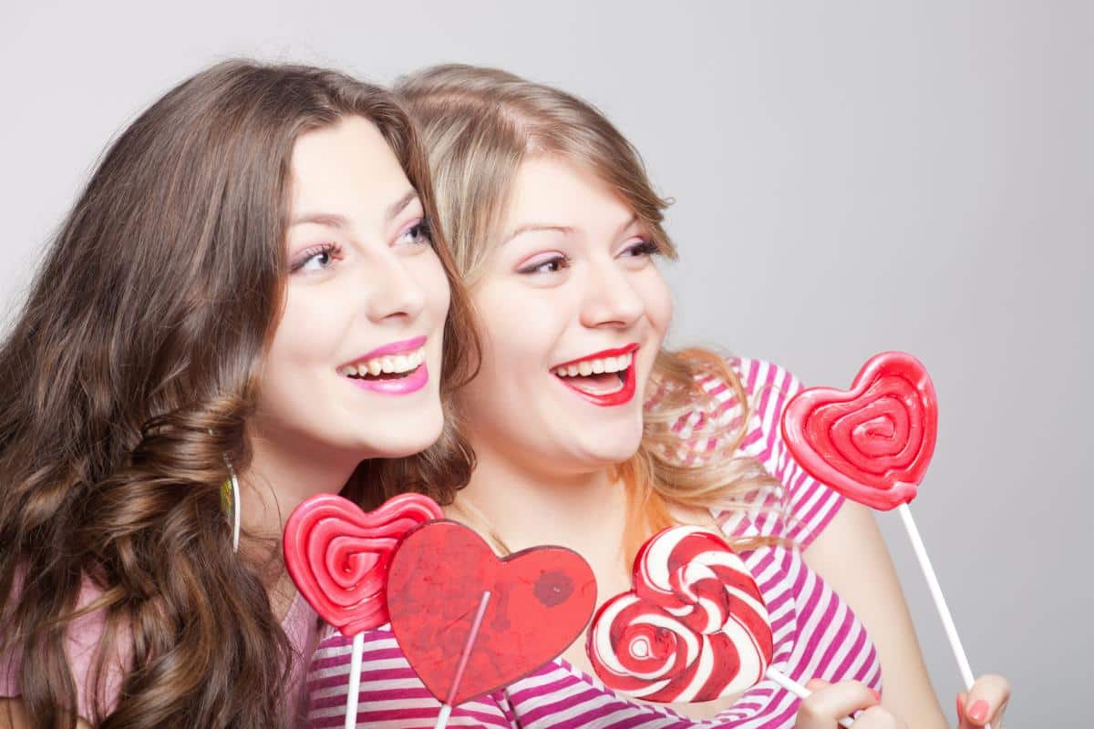 Two smiling women holding heart-shaped lollipops against white background for dental health promotion.