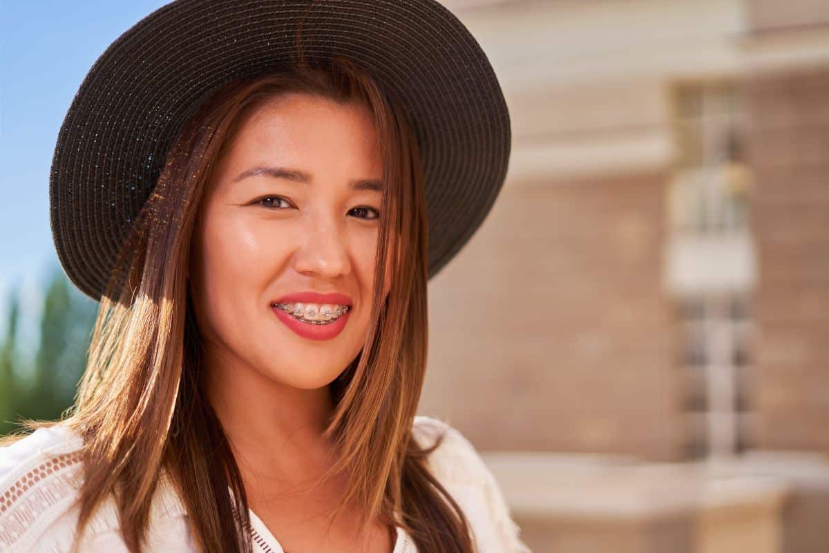 Summer Vacation with Braces Woman smiling outdoors wearing black hat and white shirt, brick building background.