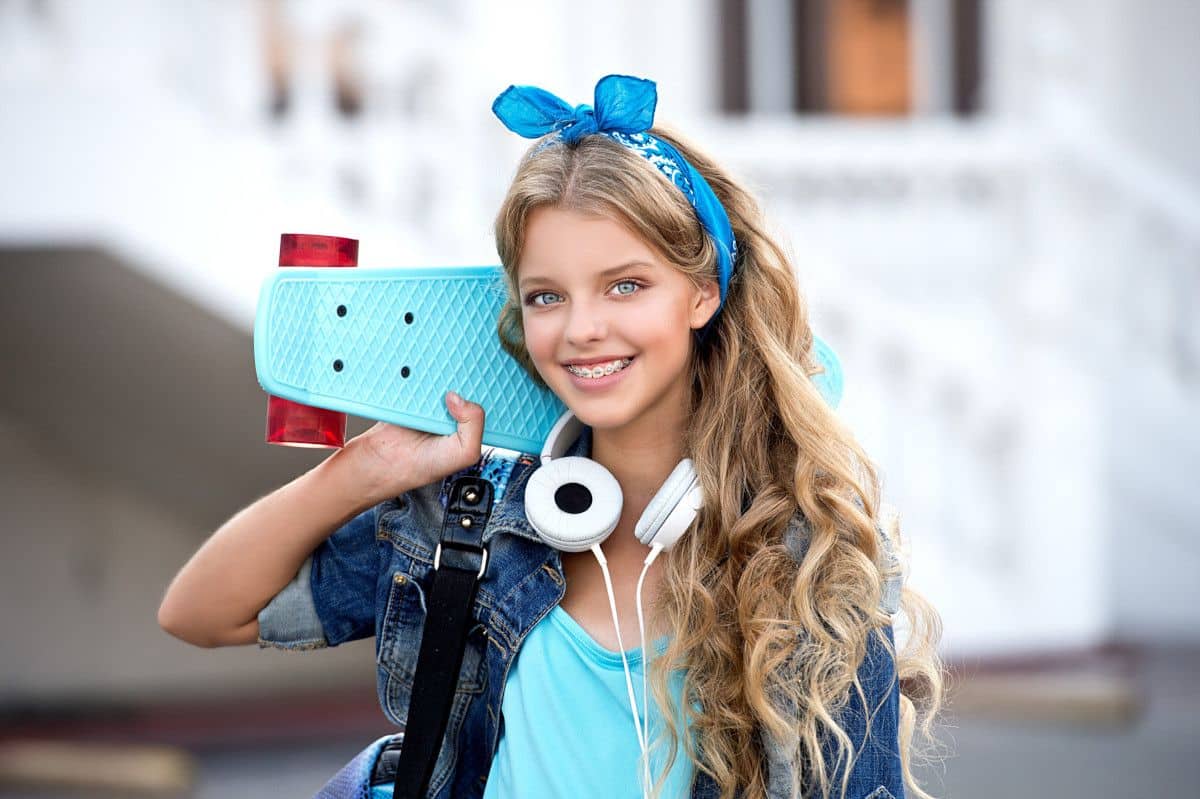 Back-to-School Braces Color Ideas Young girl holding blue toy camera with headphones around neck, smiling at camera indoors.