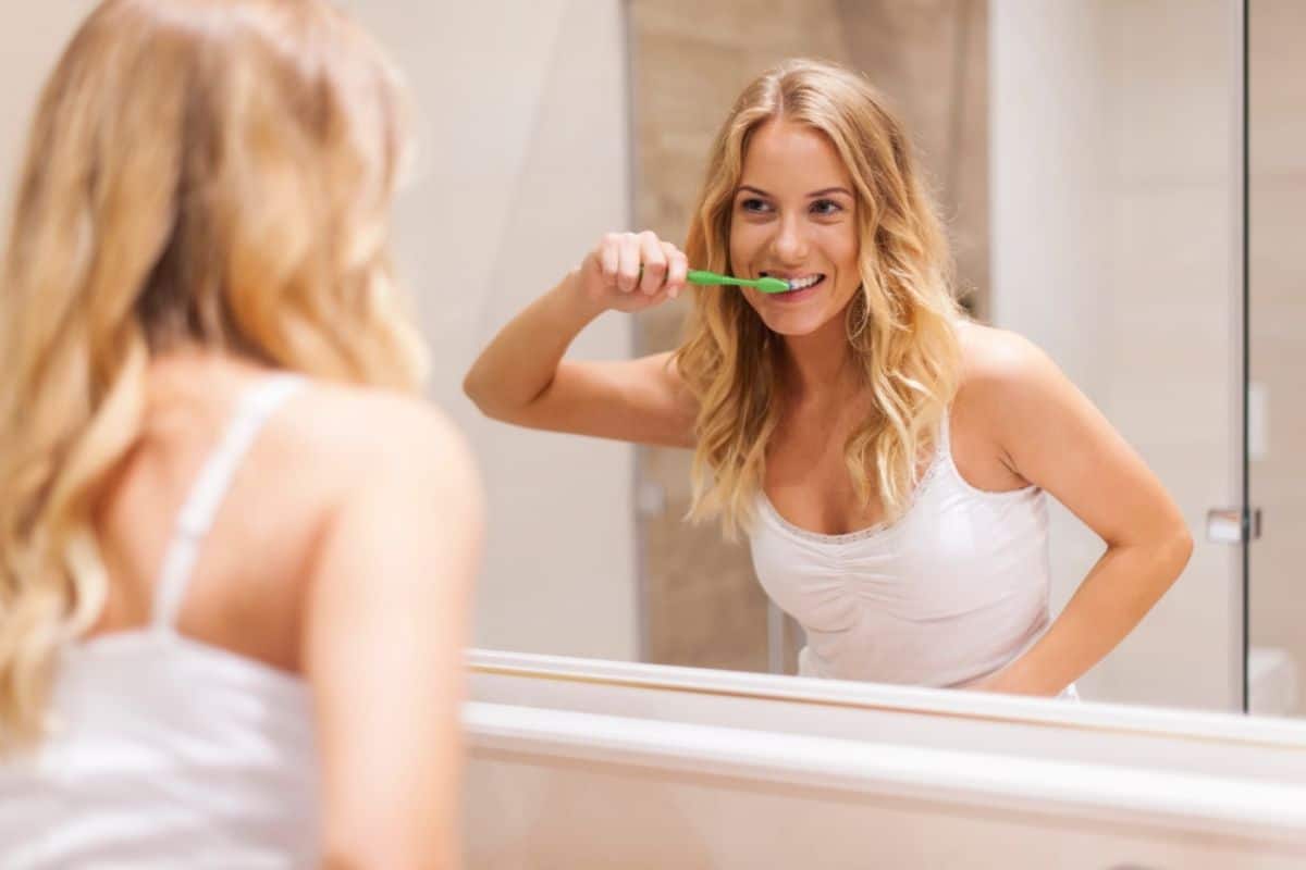 Person brushing teeth in front of bathroom mirror with a smile.